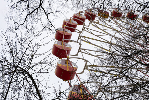 Perspective view of a large red ferris wheel behind leafless tree branches against a gray, cloudy sky.