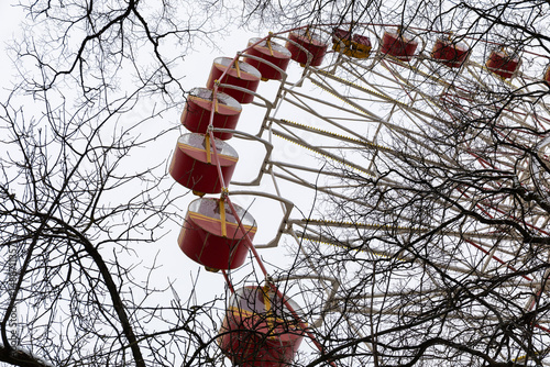 Perspective view of a large red ferris wheel behind leafless tree branches against a gray, cloudy sky.