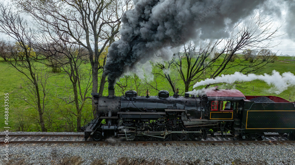Obraz premium A classic steam locomotive travels through lush green fields, releasing thick black smoke into the clear sky. It is a vibrant spring afternoon, enhancing the charming landscape.