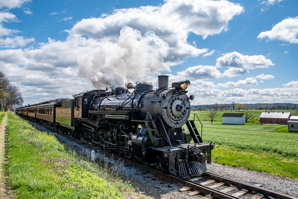 Naklejka premium A powerful steam engine makes its way along a rural track, releasing clouds of steam into the bright blue sky. Fields stretch out on either side, dotted with white barns.