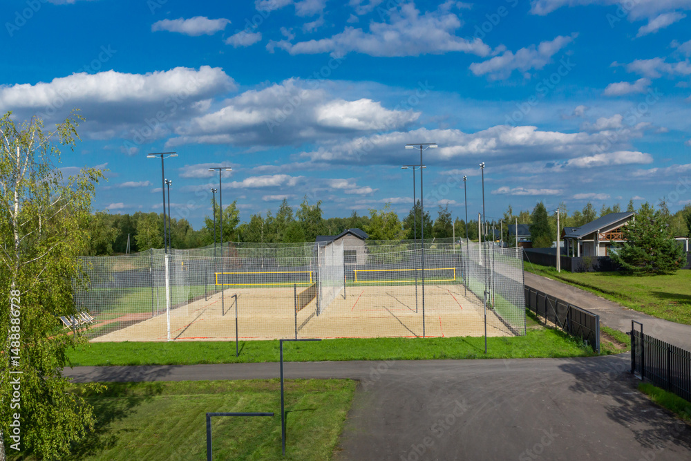 Obraz premium A spacious outdoor sports court with sandy flooring is surrounded by a mesh fence, featuring bright lighting poles. Lush green trees and houses are seen in the background under a sunny sky.