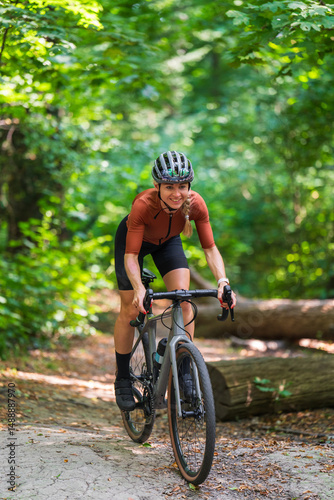 Smiling female cyclist wearing sports outfit and helmet rides gravel bike along forest trail. Concept of outdoor adventure, cycling through nature, and healthy, active lifestyle.