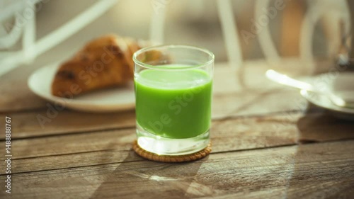 A green beverage sits in a clear glass on a wooden table, next to a plate with a flaky croissant. This leisurely snack moment occurs during a sunny afternoon, evoking a relaxed atmosphere