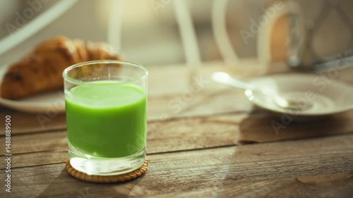 A refreshing green smoothie sits next to a croissant on a wooden table in a cozy cafe during the morning. The sunlight filters through the window, creating a warm atmosphere for breakfast