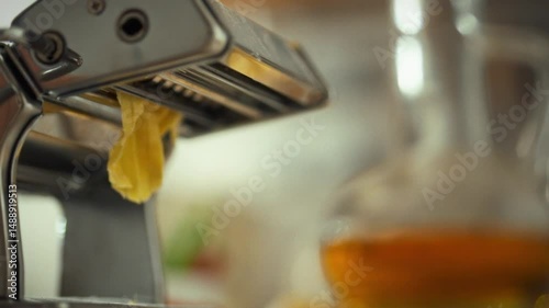 A person is using a manual pasta machine to roll out fresh pasta dough in a cozy kitchen. The soft yellow sheets of pasta are emerging, showcasing the art of homemade cooking