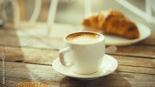 A cup of coffee sits on a white saucer beside a freshly baked croissant. Sunlight streams through a window, casting warm light on the wooden table in the cozy cafe