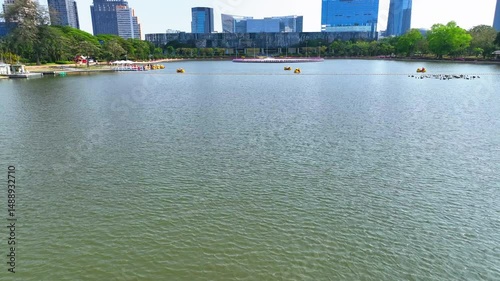 Aerial: Drone Forward Shot Of People Riding Duck Boats On Rippled Lake In Modern City Against Clear Sky - Bangkok, Thailand