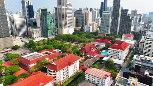 Aerial Backward Shot Of Roofed Houses Amidst Tall Modern Buildings Against Clear Sky - Bangkok, Thailand