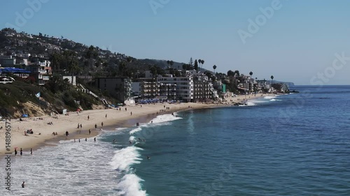Aerial: Drone Panning Shot Of Male And Female Tourists At Beach On Vacation By Buildings Over Hills In City During Sunny Day - Laguna Beach, California