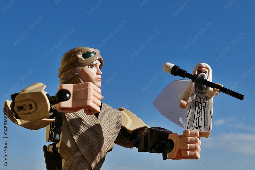 Fototapeta premium LEGO Star Wars action figure of female jedi Rey Skywalker in scavenger outfit aiming to punch small figure of LEGO Saruman from Lord Of The Rings movie with her right fist. Blue skies background.