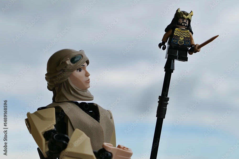Fototapeta premium LEGO Star Wars action figure of Rey Skywalker in scavenger outfit with helmet and sunglasses, holding LEGO small figure of Thorin Oakenshield from Hobit movie on tip of pole. Cloudy skies background.