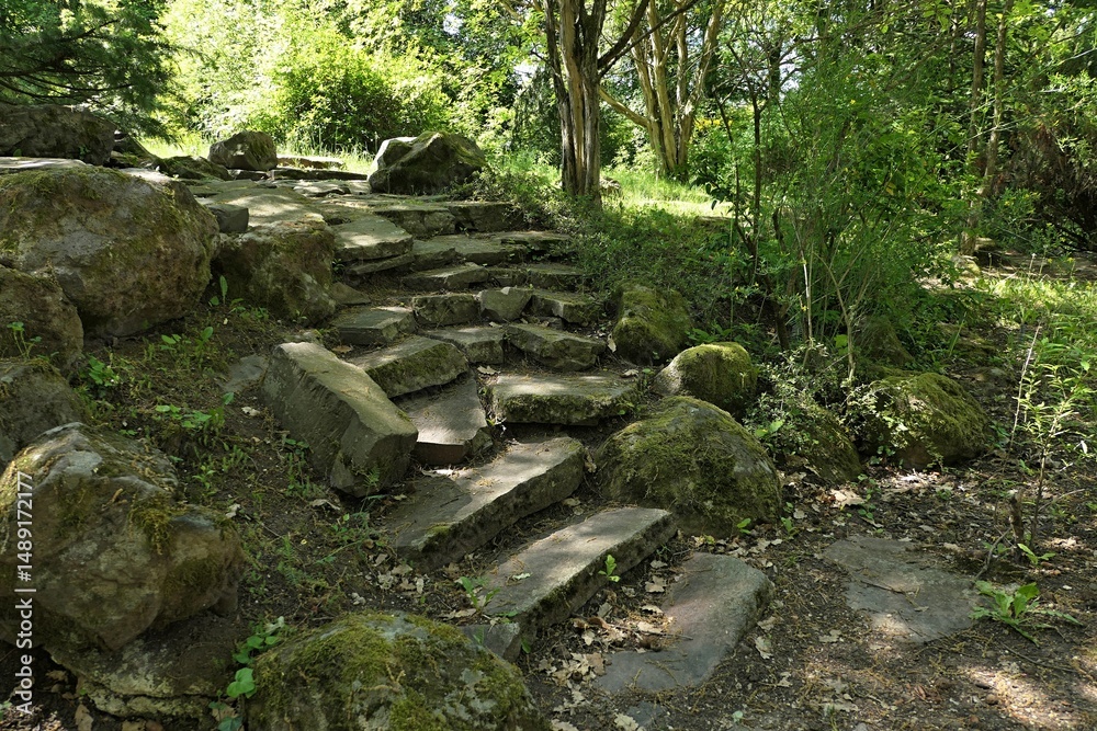 Fototapeta premium Old partially damaged stone stairs with mossy rocks around, hidden in shadow of park trees, spring daylight sunshine. 