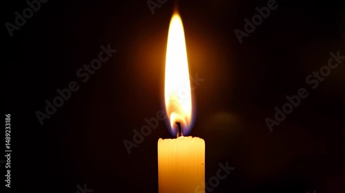 Close-up view of a single burning candle with a golden flickering flame on a black background, providing light in the darkness.