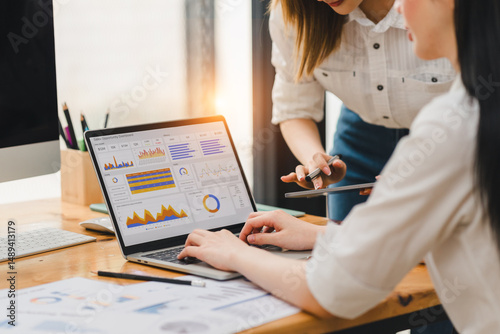 Two women working together on data analysis using laptop with colorful charts and graphs on screen, discussing business strategy in modern office environment