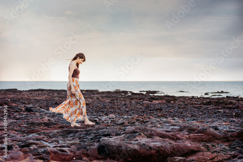 A woman wearing a long skirt walking on a rocky shore with ocean as the background
