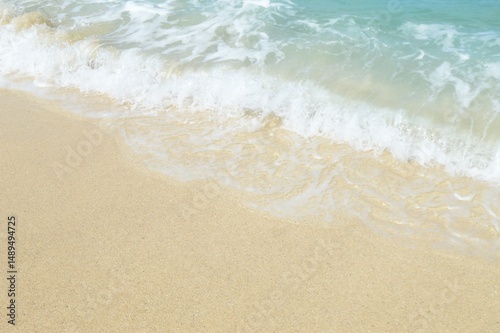 A close-up shot of a sandy beach meeting the clear turquoise ocean. Gentle waves are breaking on the shore, creating a peaceful and inviting atmosphere. The image captures the beauty of the coast