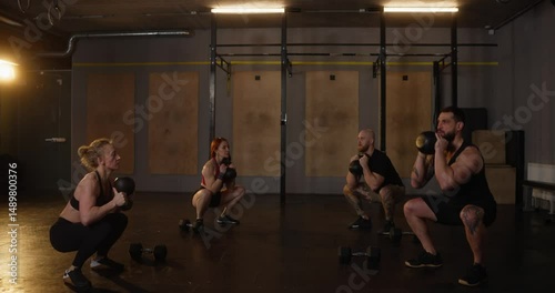 Four athletic people doing kettlebell squats and goblet squats during exercise in cozy modern fitness room. Group training warm-up the guys and girls in the gym