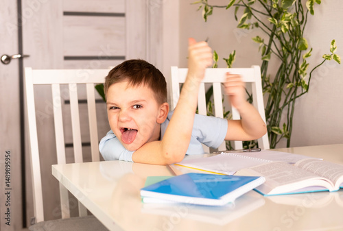 Kind of silly little boy sticking his tongue out at camera, making funny faces. Worried, not ready for test or exam, tired, upset boy doing homework in kitchen at home. Sad and angry child.