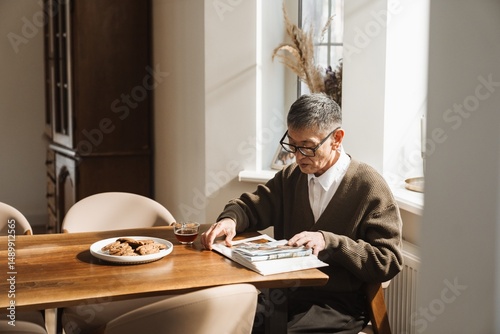An elderly Asian man looks through a family photo album while sitting at a table with tea and cookies in a cozy home atmosphere. A concept of life values, relatives, and memories.