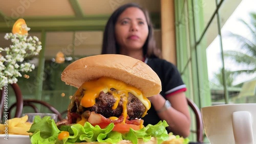 young woman drinking milkshake against background of huge juicy burger and sauces on table in restaurant
