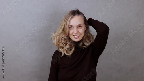 Young woman on gray background adjusting blonde hair, smiling and looking at camera, copy space, close up