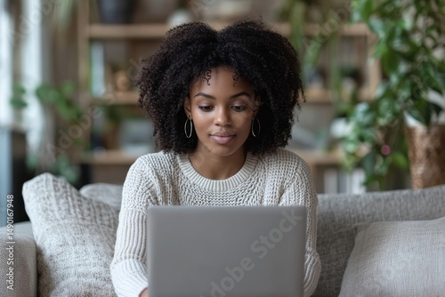 Positive black woman student having online lesson with tutor teacher, sitting on sofa and looking at laptop screen, taking notes in notepad