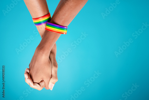 Gay couple with rainbow striped wristbands holding hands when standing against blue background with copy space