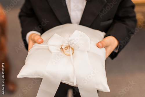 Canvas-taulu a ring bearer boy in a black suit holding a white pillow with wedding gold band