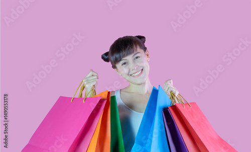 Young woman cheerful wearing eyeglasses  enjoying while holding colorful shopping bags. Lovely lady hair two buns wearing red eyewear hold shopping bags isolated on pink color background.