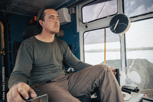 Crane operator controls the joystick of the crane, front side view inside the cabin. Man operates crane at loading dock on river for coal barge transport while managing port logistics during day shift