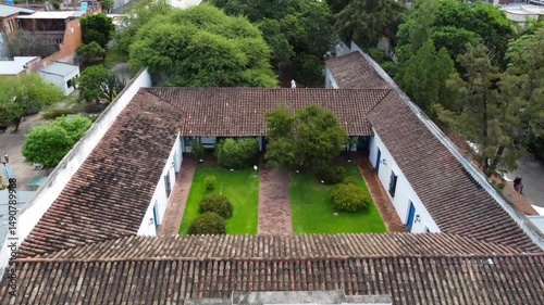 Drone flight over the colonial facade of the Casa Histórica de Tucumán, its windows, flags, shields, and the lush green vegetation of its inner courtyards, including the historic well.