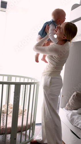 Young mother gently lifting crying baby daughter from crib in bright nursery. Tender parenting moment, bonding, care and love at home. Real indoor lifestyle scene with soft light and warmth
