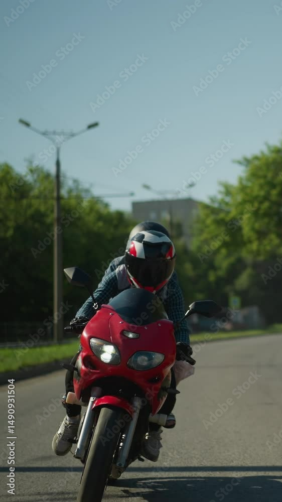 Two friends ride a red power bike, both wearing helmets, navigating the road with parked cars along the roadside, electric poles and lush trees are visible in the background Two friends ride a red power bike, both wearing helmets, navigating the road with parked cars along the roadside, electric poles and lush trees are visible in the background