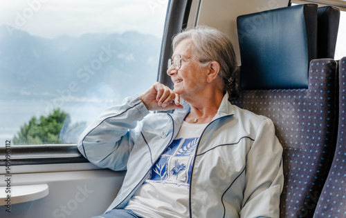Senior smiling woman with eyeglasses enjoys the train ride sitting in passenger seat by the window looking out the landscape. Happy grandmother enjoying travel