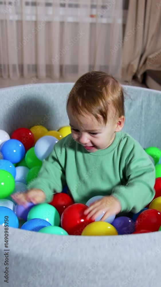 Excited baby boy clapping hands by the colorful toys in the basin he's sitting in. Lovely toddler playing indoors. Domestic cat watching the child. Vertical video Excited baby boy clapping hands by the colorful toys in the basin he's sitting in. Lovely toddler playing indoors. Domestic cat watching the child. Vertical video