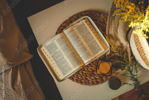 Slika na platnu Open Bible on white table with candle, linen fabric, and yellow wildflowers in s