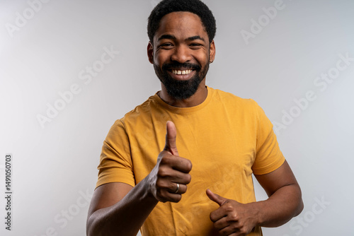 Handsome smiling Black man in a yellow t-shirt showing thumbs up. Confident positive expression, isolated on grey background. Modern casual lifestyle.