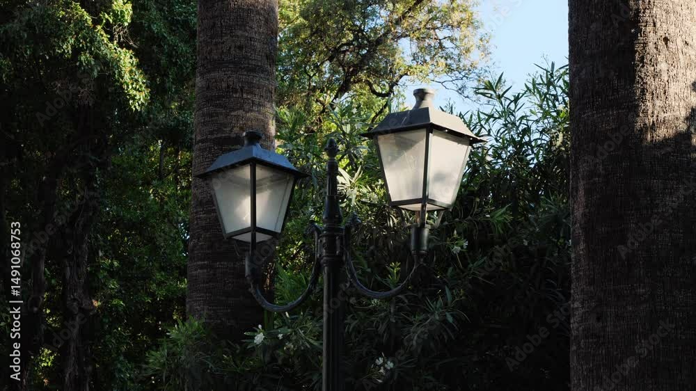 Medium shot of the upper part of a vintage double street lamp in the National Garden of Athens, Greece. The pole splits into two lanterns. Trees fill the background. No people.