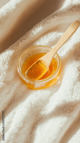Golden sugaring paste in glass jar with wooden spatula on soft white towels, natural light, spa beauty concept, clean white background
