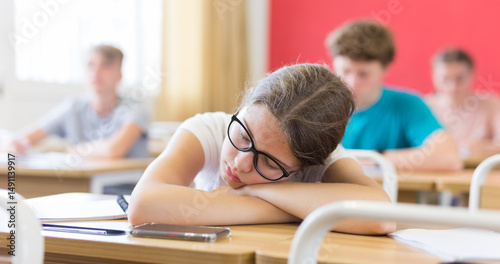 Exhausted teen girl with glasses sleeping at desk in classroom during lesson on blurred background of classmates ..