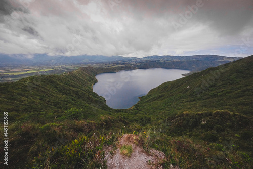 The image shows Cuicocha Lake, a stunning crater lake in Ecuador, nestled within a volcanic caldera. Deep blue waters reflect the sky, surrounded by steep, green hills and distant mountains under a