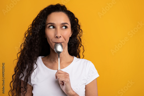 Portrait of excited hungry young woman holding spoon in her mouth dreaming about delicious healthy food, choosing something to eat looking aside at free copy space, yellow orange studio background