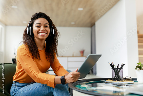 People And Technology. Portrait of young smiling black woman in wireless headset holding using digital tablet sitting on chair in living room, looking posing at camera, free copy space, banner photo