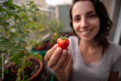 Close-up of woman's hand holding small red tomato, blurred background shows garden and smiling woman, showcasing homegrown produce and healthy lifestyle