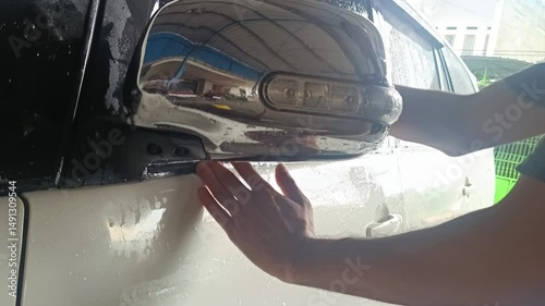 Close-up shot of a hand washing a car side mirror with soap, ensuring cleanliness, luster and care for the automobile's bodywork, maintaining its quality.