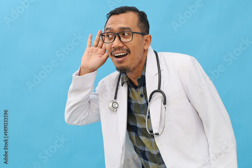 Asian adult male doctor standing isolated on blue background, wearing doctor's coat, stethoscope hanging on neck, listening to something with hand on ear, glancing sideways.