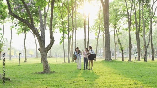 Group of Asian school girls discussing learning about researching academic content using tablets, taking notes in notebooks under a tree in a park.