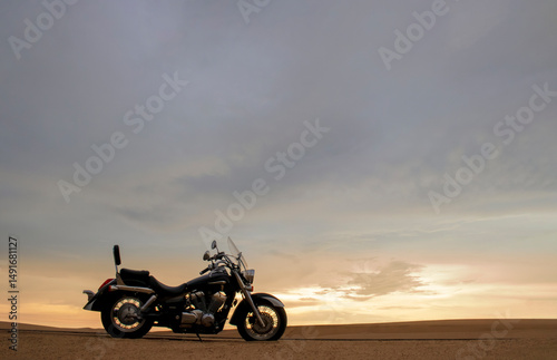 A lone motorcycle stands on the sandy surface of the Namib Desert against a dramatic cloudy sky. The sunset lighting highlights the contrast between nature and machine.