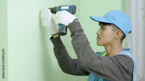 Asian male worker uses a cordless drill as a screwdriver to fasten screws on a gypsum board wall.