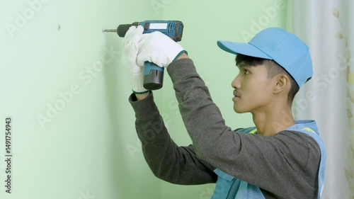 Asian male worker uses a cordless drill as a screwdriver to fasten screws on a gypsum board wall.